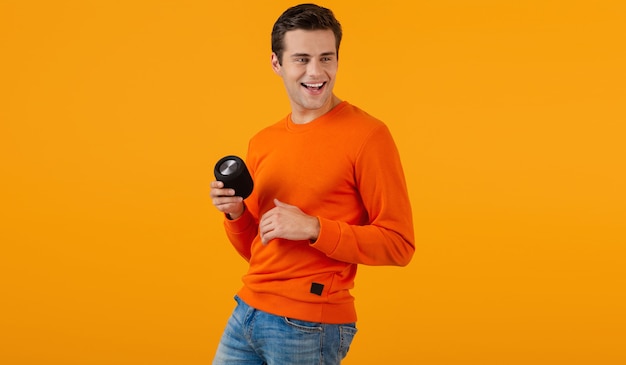 Stijlvolle lachende jonge man in oranje trui met draadloze luidspreker gelukkig luisteren naar muziek plezier op oranje