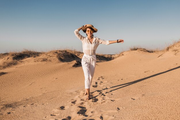 Stijlvolle lachende mooie gelukkige vrouw rennen en springen in woestijnzand in witte outfit met strooien hoed op zonsondergang