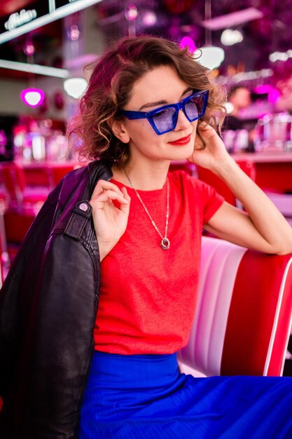 Stijlvolle lachende vrouw in kleurrijke outfit in retro vintage jaren '50 café zittend aan tafel met jas blauwe rok en rode shirt zonnebril plezier in vrolijke stemming