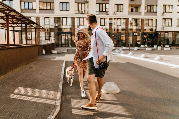 Stijlvolle man en vrouw in zomeroutfits rennen en spelen met hun hond op de achtergrond van het appartementencomplex. Man in licht overhemd houdt zijn geliefde hand vast en draagt de camera.