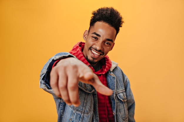 Stijlvolle man in een rood sweatshirt wijst met zijn vinger naar de camera en poseert op een geïsoleerde muur