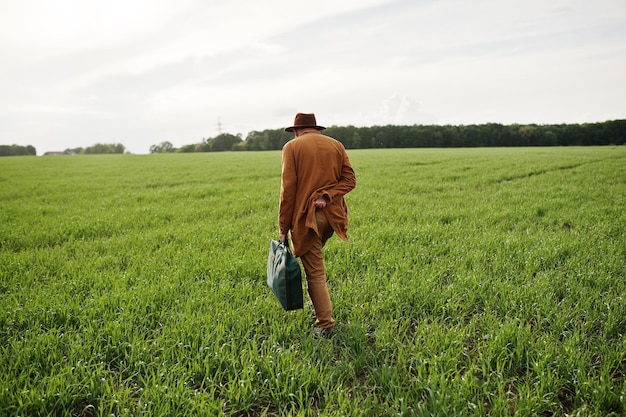 Stijlvolle man in glazen bruine jas en hoed met tas op groen veld