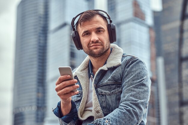 Stijlvolle man met spijkerjasje die naar muziek luistert en smartphone gebruikt die op bewolkte ochtend voor wolkenkrabbers in de stad Moskou zit.