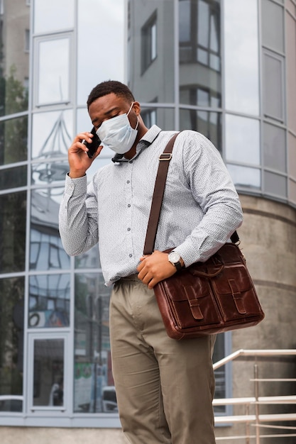 Gratis foto stijlvolle man praten aan de telefoon met masker op terwijl hij onderweg is naar zijn werk tijdens pandemie