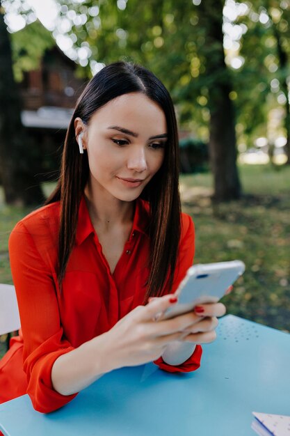 Stijlvolle meisje met donker haar in rode blouse is poseren met telefoon en kopje koffie op de achtergrond van groen park Aantrekkelijke vrouw in stijlvolle outfit glimlachend buiten