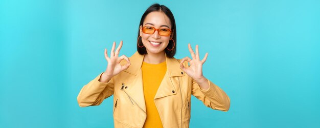 Stijlvolle, mooie aziatische vrouw met een zonnebril die verbaasd glimlacht en een goed teken toont dat iets aanbeveelt