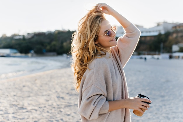 Stijlvolle mooie vrouw blinde in beige oversized trui en bruine zonnebril loopt aling strand met kartonnen kopje thee.