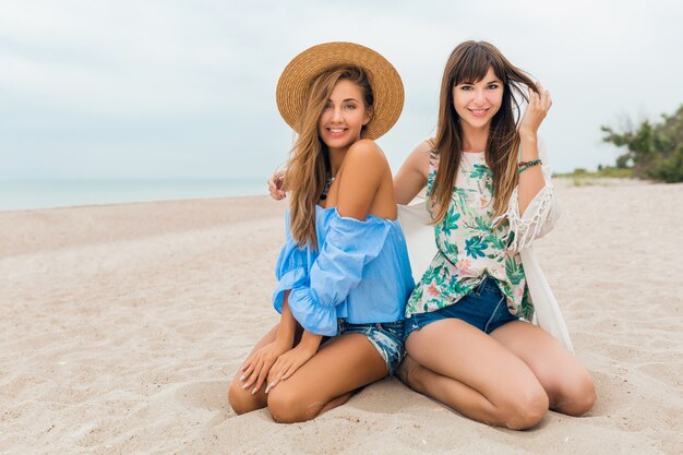 Stijlvolle mooie vrouwen op zomervakantie op tropisch strand, bohemien stijl, vrienden reizen samen, modetrend, accessoires, glimlachen, vrolijke emotie, positieve stemming, strooien hoed, zittend op zand