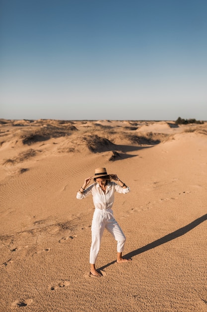 Stijlvolle mooie zorgeloze gelukkige vrouw wandelen in woestijnzand gekleed in witte broek en blouse met strooien hoed op zonsondergang