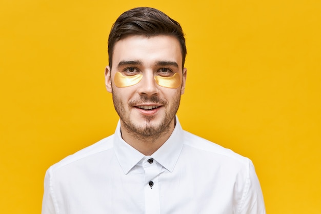 Stijlvolle ongeschoren jonge blanke man die een oogmasker draagt om uitdroging en donkere kringen aan te pakken vanwege een stressvolle levensstijl, poseren tegen een gele muur,