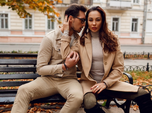 Stijlvolle paar verliefd poseren buiten. herfst modetrends. brunette model met stijlvolle man in beige jas zittend op de bank.