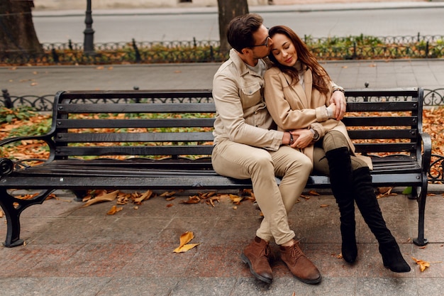 Stijlvolle paar verliefd poseren buiten. Herfst modetrends. Brunette model met stijlvolle man in beige jas zittend op de bank.