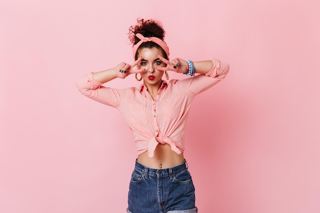 Gratis foto stijlvolle pinup meisje met roze hoofdband vredestekens tonen portret van vrouw in roze blouse en korte broek op geïsoleerde background