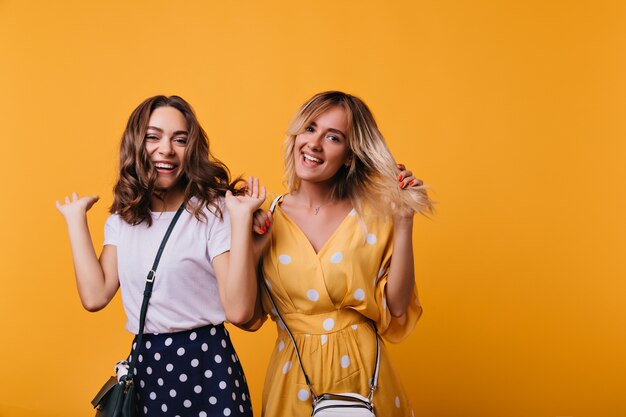 Stijlvolle schattige dames dansen en lachen in hun vrije tijd. Geweldige vrouwelijke modellen die geluk uitdrukken terwijl ze poseren op oranje.