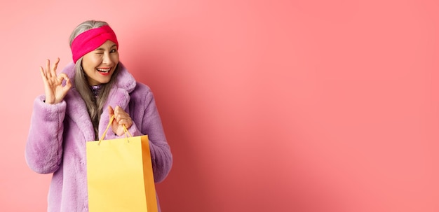 Gratis foto stijlvolle senior aziatische vrouw die winkel aanbeveelt die boodschappentas vasthoudt en knipoogt met ok teken glimlachen