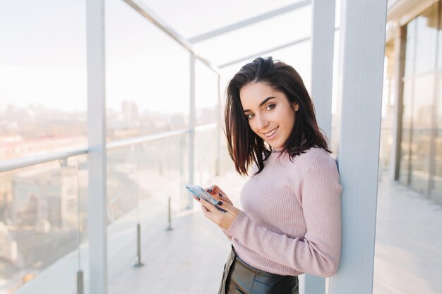 Stijlvolle stad portret jonge modieuze brunette vrouw met behulp van telefoon op terras op uitzicht op de stad. Aantrekkelijke zakenvrouw, vrolijke sfeer, glimlachend.