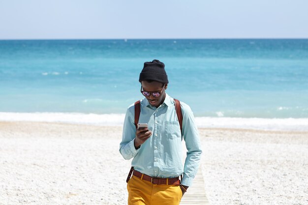 Stijlvolle student met een donkere huidskleurige knapzak die trendy kleding draagt die op de promenade staat na een ochtendwandeling op het woestijnstrand