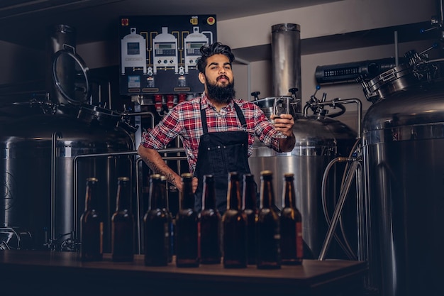 Stijlvolle volle bebaarde Indiase man in een fleece shirt en schort houdt een glas bier vast, staande achter de toonbank in een brouwerij.