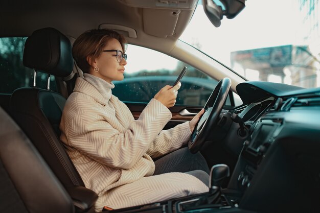 stijlvolle vrouw die in de auto zit, gekleed in winterjasstijl en bril met smartphone