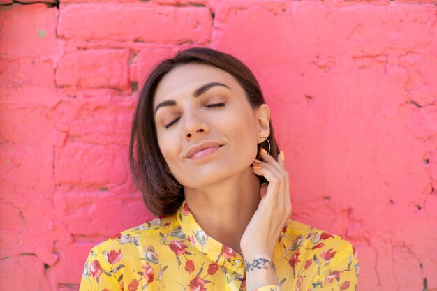 Stijlvolle vrouw in gele zomerjurk op roze bakstenen muur gelukkig kalm en positief