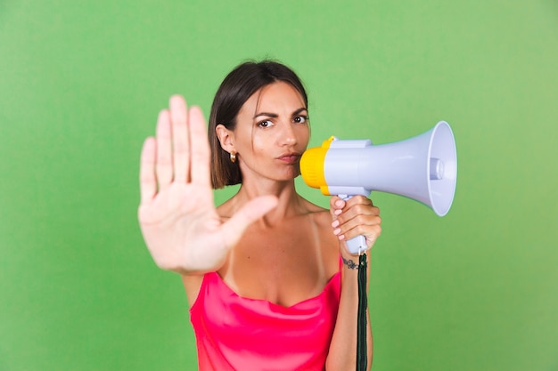 Stijlvolle vrouw in roze zijden jurk op groen, met megafoon, geïsoleerd, stop gebaar met serieus gezicht