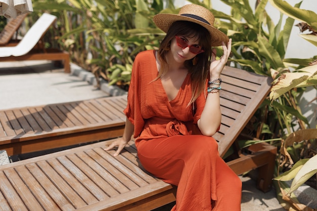Stijlvolle vrouw in strohoed en oranje playsuit rustend op haar villa tijdens vakantie op Bali.