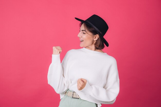 Stijlvolle vrouw in witte casual trui en hoed op rood roze muur