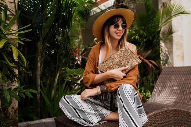 Stijlvolle vrouw in zomerkleren ontspannen in hotel en genieten van trendy zonnebril, strooien hoed en handtas, boheemse armbanden en accessoires.