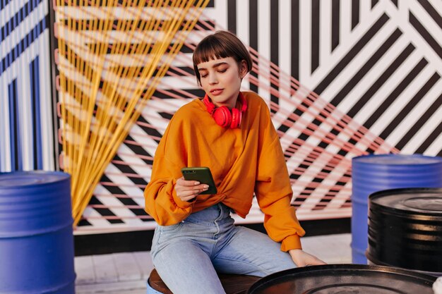 Stijlvolle vrouw met kort haar in oranje outfit die buiten zit Charmant meisje in rode koptelefoon en spijkerbroek met telefoon op gestreepte achtergrond