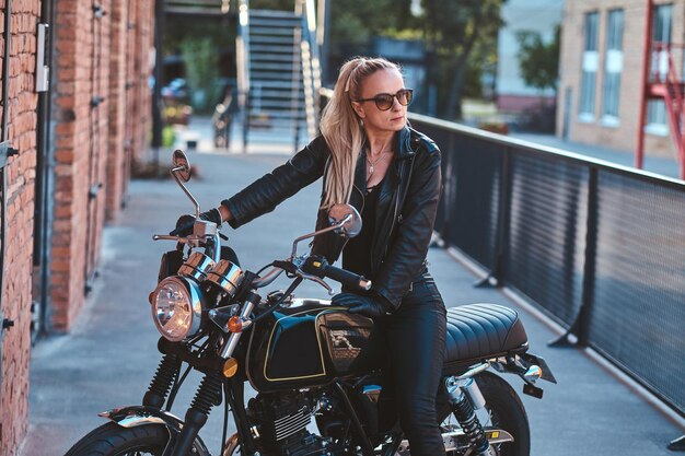 Stijlvolle vrouwelijke motorrijder zit op haar zwarte fiets terwijl ze poseert voor een fotoshoot op straat.