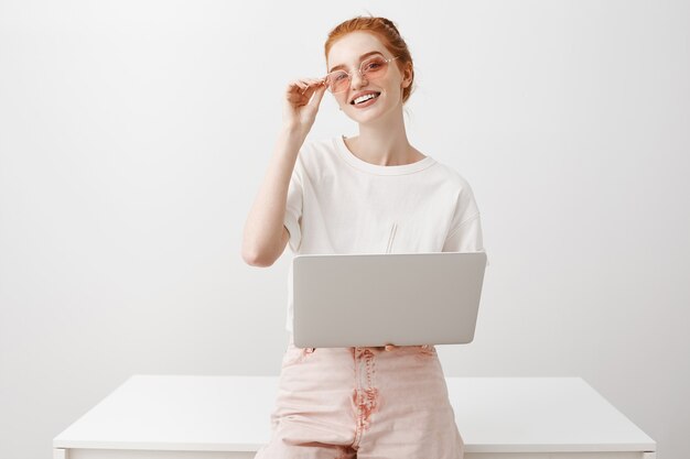 Stijlvolle vrouwelijke roodharige in zonnebril die met laptop werkt