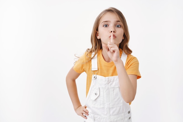 Stil alsjeblieft. Vastberaden gehoorzaam schattig blond meisje dat een stil teken toont, stilzwijgen vraagt om stilte, vinger op de lippen houdt, luide muziek uitzet, geheim deelt, op witte muur staat