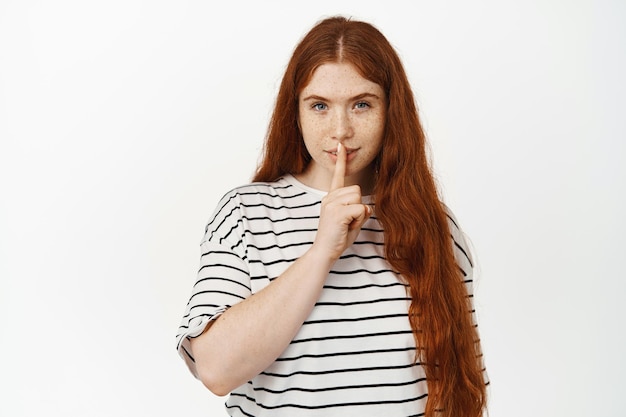 Stil, vertel het aan niemand. Mysterieus roodharig meisje zwijgt, maakt shh-teken met de vinger op de lippen en glimlacht, vertelt geheim, vraagt om stil te blijven, staande tegen een witte achtergrond