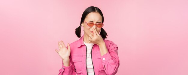 Stinkend Aziatisch meisje met zonnebril en stijlvolle outfit sluit haar neus van walging, houdt niet van slechte geur die over roze achtergrond staat