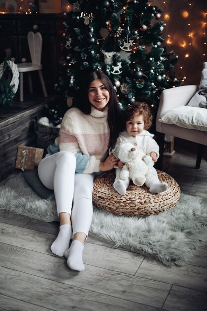 Stock foto van glimlachende aantrekkelijke moeder en baby zitten onder versierde kerstboom thuis.