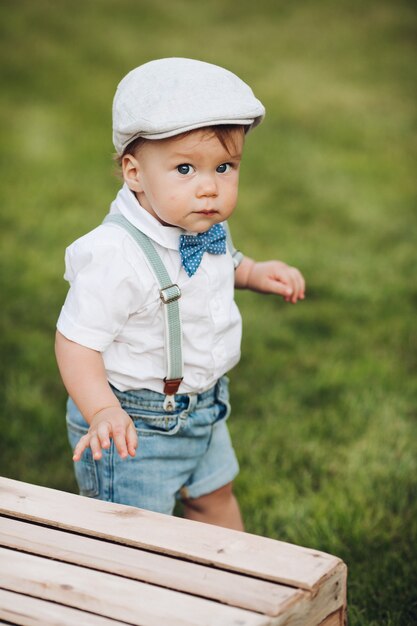 Stockfoto portret van een schatje in hoed, shirt, korte broek en strik met bretels camera kijken tijdens het wandelen op groen gazon in de achtertuin. Kijkend naar de camera met nieuwsgierigheid.