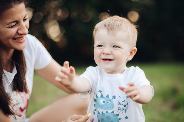 Stockfoto van een mooie babyjongen in t-shirt die de eerste stappen in het park zet met zijn moeder ernaast. Bokeh. Onscherpe achtergrond. Kleine jongen strekt zijn armen uit tijdens het lopen.