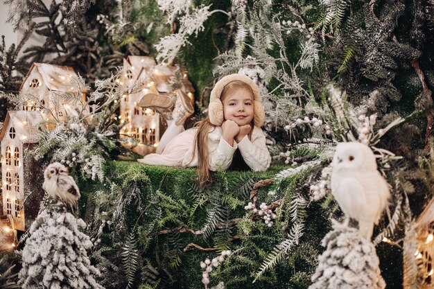 Stockfoto van een schattig klein blank kind in beige oorbeschermers met kin op handen omringd door kerstversieringen en glimlachend in de camera. Winterwonderland-concept.