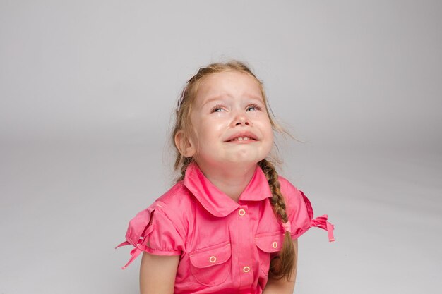 Stockfoto van lief klein meisje met vlechten in roze jurk huilend zittend op de vloer met blote voeten Ze kijkt naar de camera terwijl ze snikt