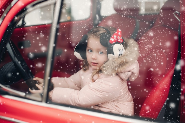 Stockfoto van schattig klein meisje in mickey oorbeschermers en winterjas zittend op de bestuurdersstoel in rode auto. ze kijkt naar de camera door het raam onder de sneeuwval.