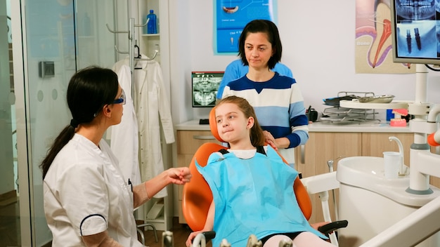 Stomatoloog legt aan klein meisje het reinigingsproces van tanden uit terwijl man-assistent gesteriliseerde hulpmiddelen voorbereidt voor onderzoek. Verpleegkundige en arts werken samen in moderne stomatologische kliniek
