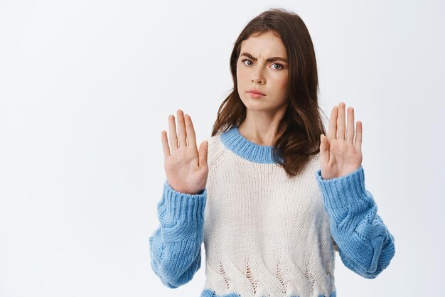 Stop nu Ernstige fronsende vrouw die handen opsteekt met blokgebaar dat nee zegt en een slecht aanbod weigert in trui tegen witte achtergrond