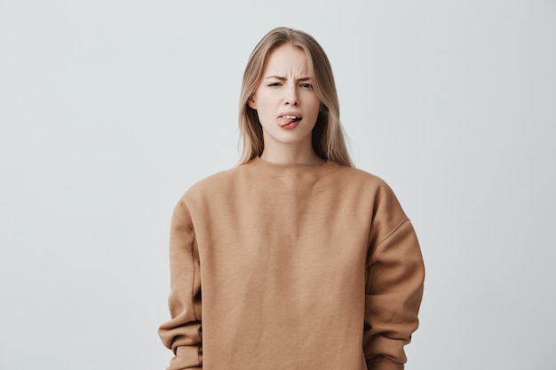 Stoute mooie vrouw met lang blond haar in beige trui die zich misdraagt, tong uitsteken als een teken van ongehoorzaamheid, protest en gebrek aan respect. Emoties, reactie, gevoelens en houding