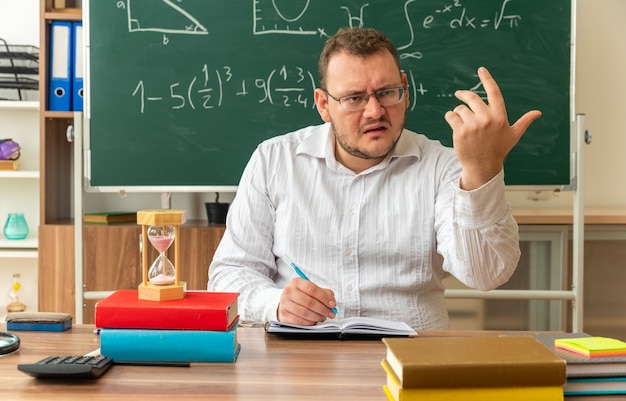 Strenge jonge blonde leraar met een bril die aan het bureau zit met schoolhulpmiddelen in de klas met een pen die naar de zijkant kijkt, kom hier gebaar