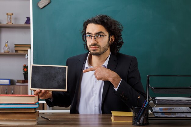 Strenge mannelijke leraar met een bril die vasthoudt en wijst naar een mini-bord dat aan tafel zit met schoolhulpmiddelen in de klas