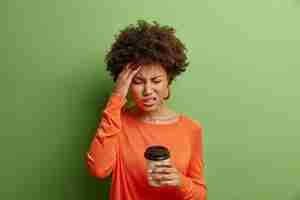 Gratis foto stressvolle afro-amerikaanse vrouw voelt vreselijke hoofdpijn grijnst van pijn sluit ogen raakt tempel drinkt koffie draagt casual oranje trui geïsoleerd over levendige groene muur