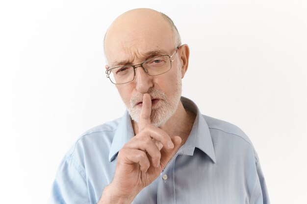 Strikt geïrriteerde bejaarde CEO in blauw shirt en bril die tijdens de conferentie een zwijgend teken maakte en vroeg om zachtjes te spreken. Senior man houdt zijn wijsvinger op de lippen en zegt Shh