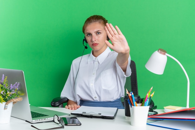 Strikt jong blond callcentermeisje met een hoofdtelefoon die aan het bureau zit met uitrustingsstukken die een stopgebaar doen