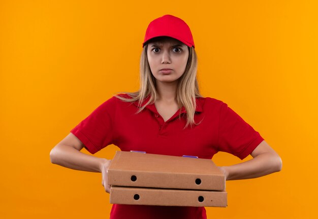 Strikt jong leveringsmeisje die rood uniform en GLB dragen die pizzadoos houden die op oranje muur wordt geïsoleerd