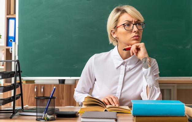 Strikte jonge blonde vrouwelijke leraar met een bril die aan een bureau zit met schoolhulpmiddelen in de klas die de hand op een open boek houdt en onder de kin met een pen